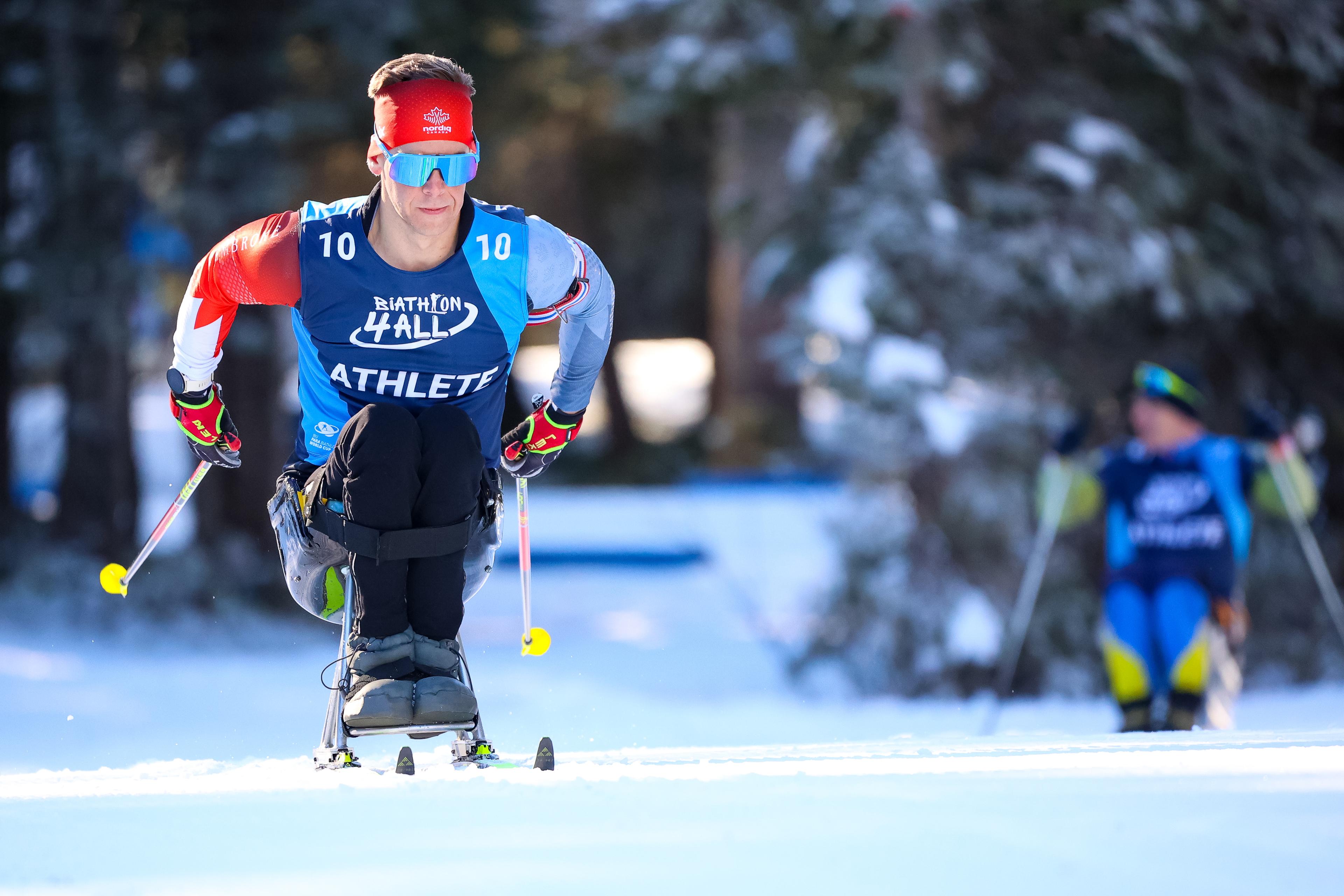 A Para biathlon athlete with bib number 10 competing in the sit-ski category on a snowy trail. The athlete wears a red headband, blue sunglasses, and a Biathlon 4 All vest while using ski poles to navigate. Another competitor is visible in the blurred background against a forest line.