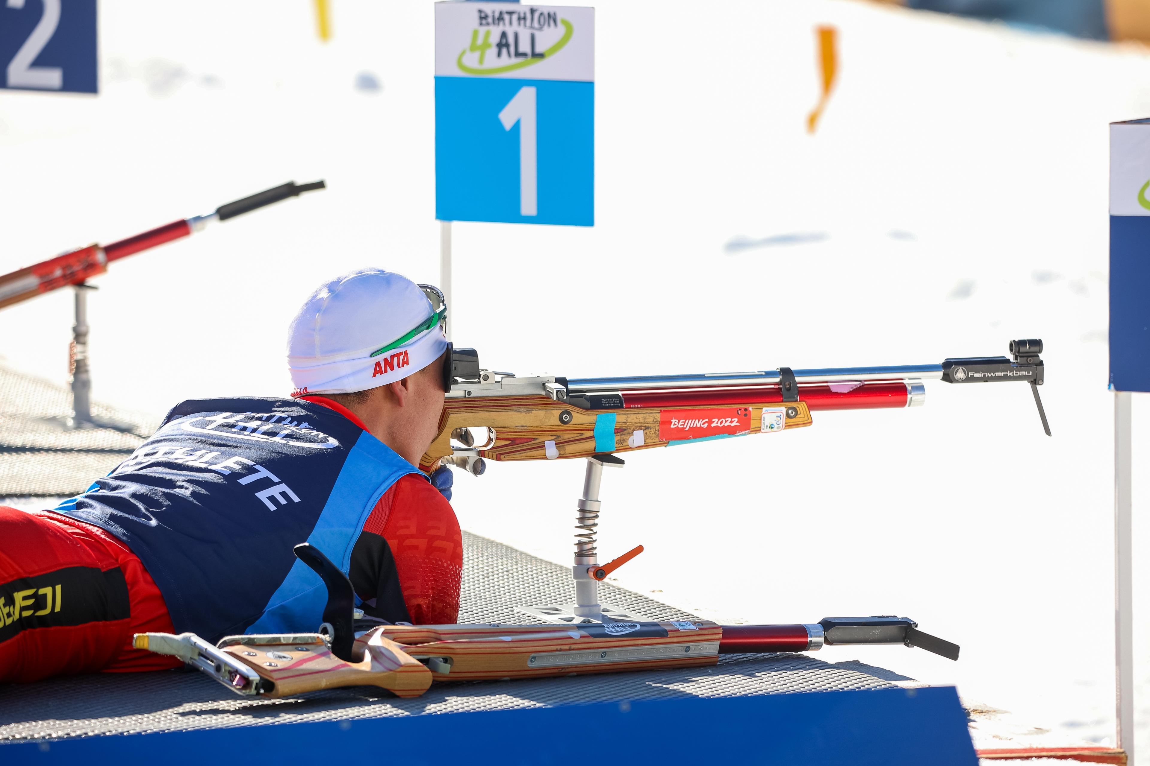 A Para biathlon athlete lies prone on a shooting mat, aiming a specialized air rifle at a target. The athlete wears a red and blue racing suit and a white ANTA branded cap. The rifle, resting on a spring-loaded stand, is wood-grained with a red barrel and a Beijing 2022 sticker.