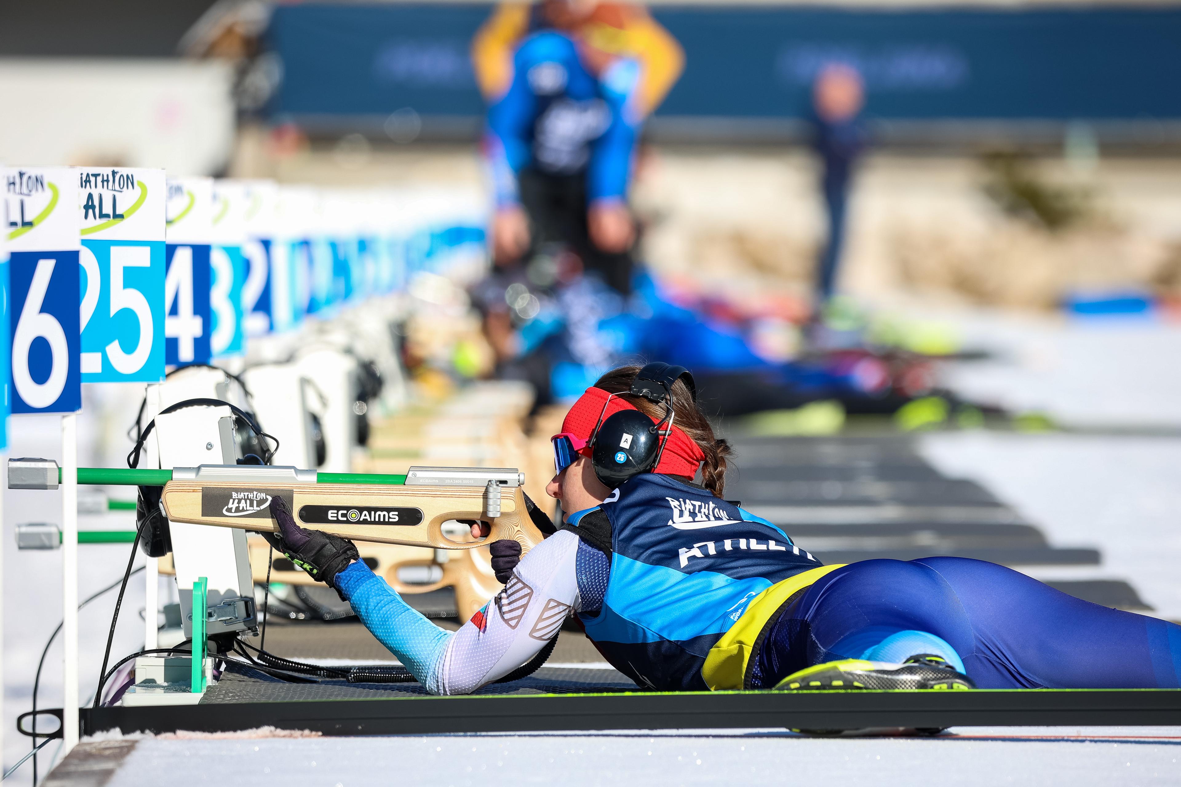 A Para biathlon athlete lies prone at a shooting range, aiming an electronic rifle. The athlete wears a red headband and black noise-canceling headphones. The background shows a line of blue and white lane markers numbered 4, 5, and 6 with the Biathlon 4 All logo.